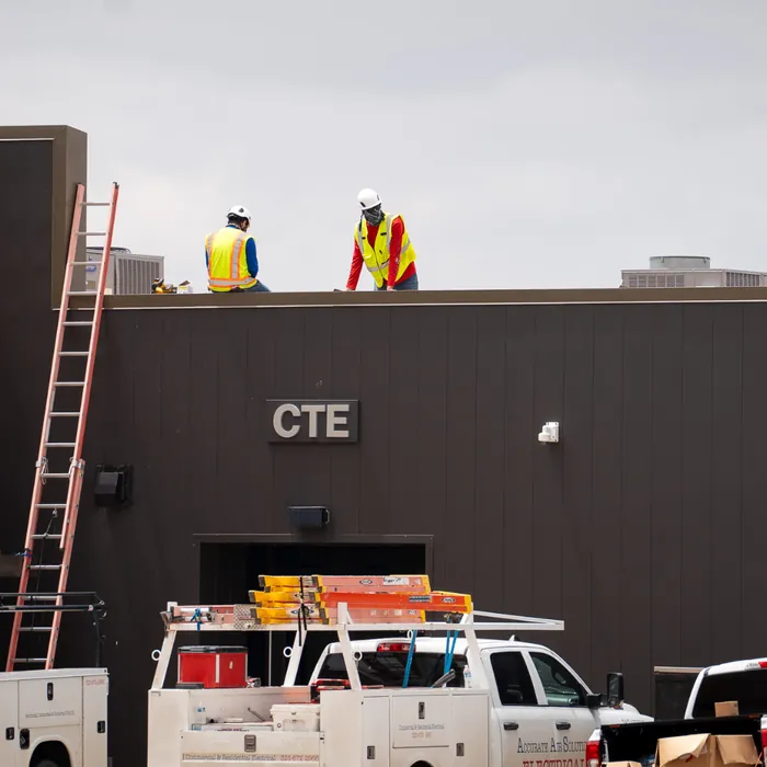 Two Accurate Air Solutions crew members in hi-vis and hard hats on the roof of a school CTE building, access ladder in place